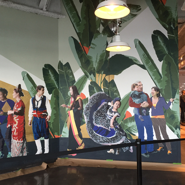 Wide installation view of mural figures in colorful dress beneath towering banana leaves and industrial pendant lights.
