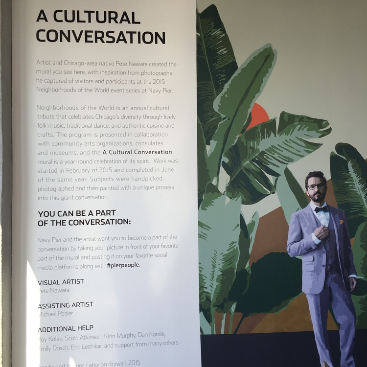 Installation photograph of a tall text panel beside a mural of large banana leaves and a man in a light suit.